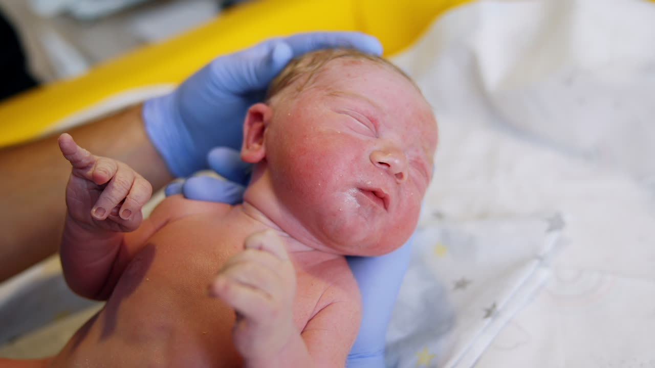Obstetrician checking up just born Caucasian baby. Medic's gloved hand touches blond-haired kid's head. Close up.