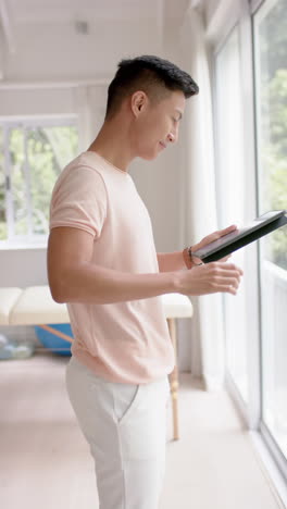 Vertical video of portrait of happy asian male physiotherapist with tablet in studio, slow motion