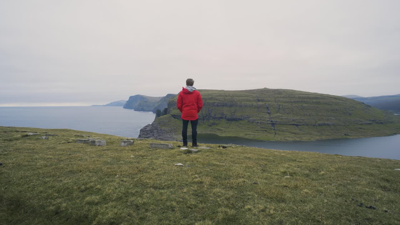 hombre con chaqueta roja parado en una colina y mirando la increíble vista del paisaje con fiordos, montañas y acantilados en un día nublado en vagar en las islas feroe