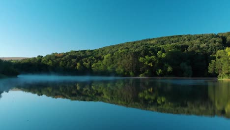 early morning low drone shot over glossy lake with some fog