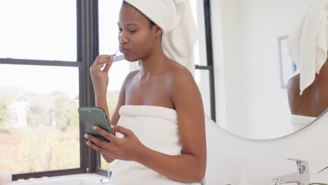 happy biracial woman with towel on head using smartphone and brushing teeth, slow motion