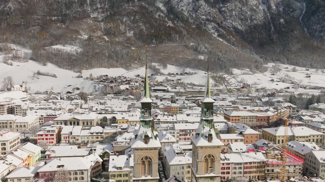 Drone flight behind the Stadtkirche in snow-covered Glarus on a clear winter day. The aerial view reveals the church towers, colorful buildings, and surrounding alpine mountains