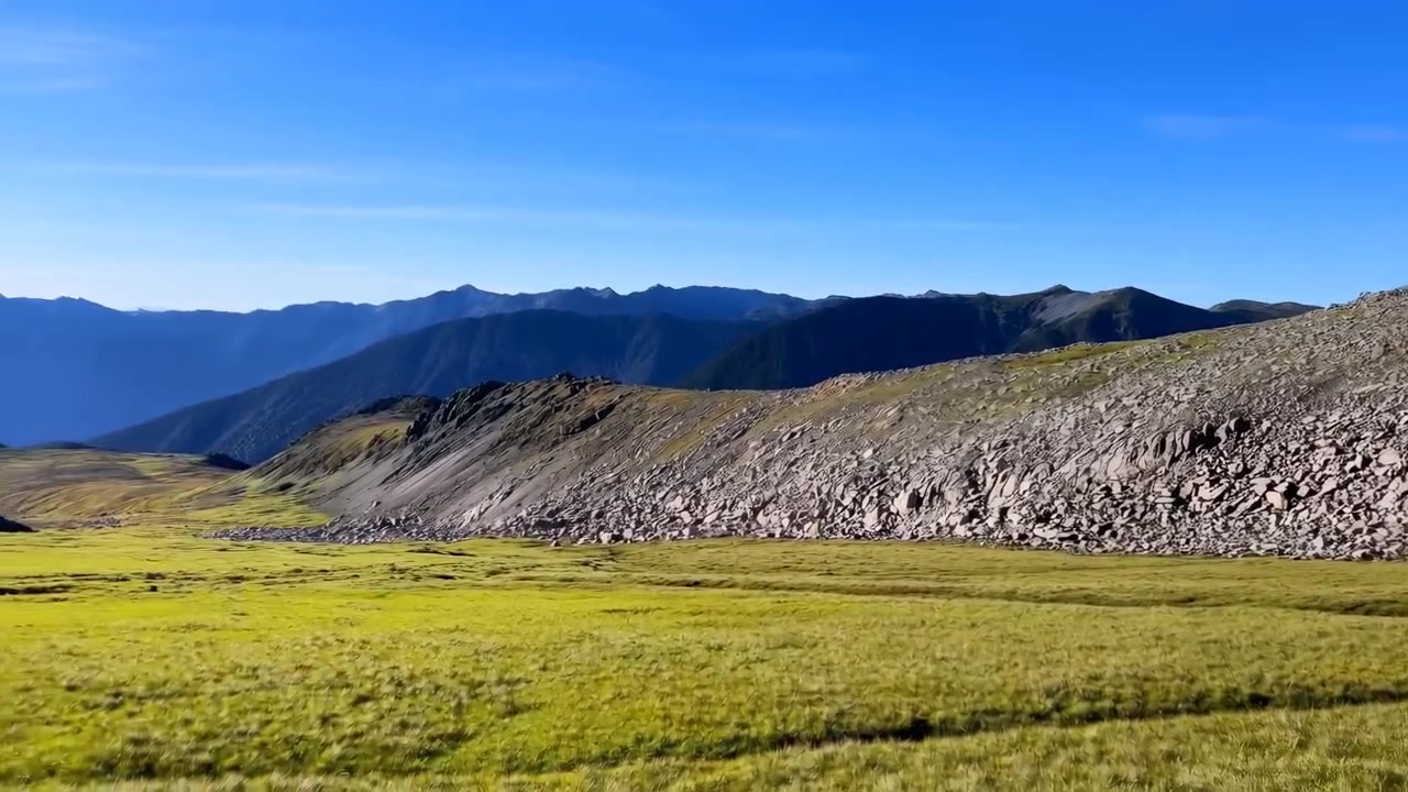 Un ampio paesaggio montano caratterizzato da un ripido ghiaione di rocce e massi spezzati che si innalza al centro-destra, con un altopiano erboso verde in primo piano