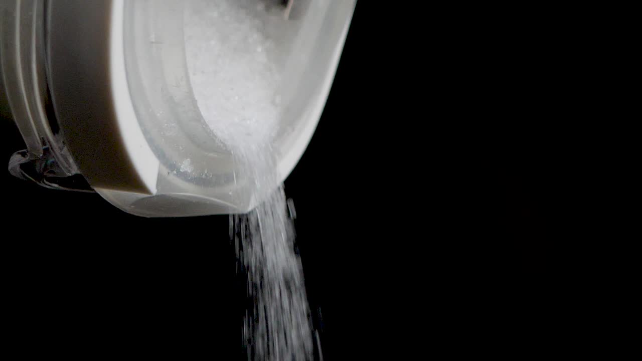 격리된 검정색 배경에서 슬로우 모션으로 투명한 용기에서 설탕을 뿌리는 샷을 닫습니다.