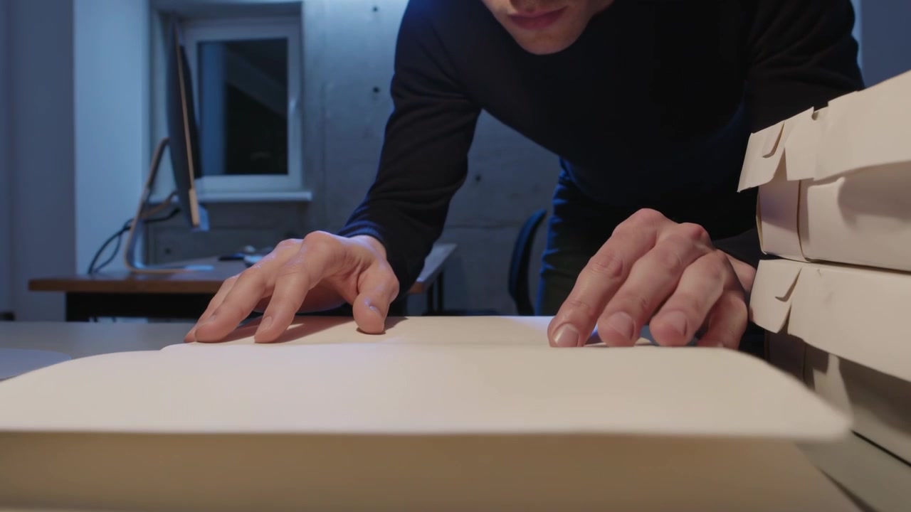 Uomo con maniche scure che posiziona un quaderno sul bordo della scrivania, sfogliando e lisciando le pagine vicino a scatole per schizzi