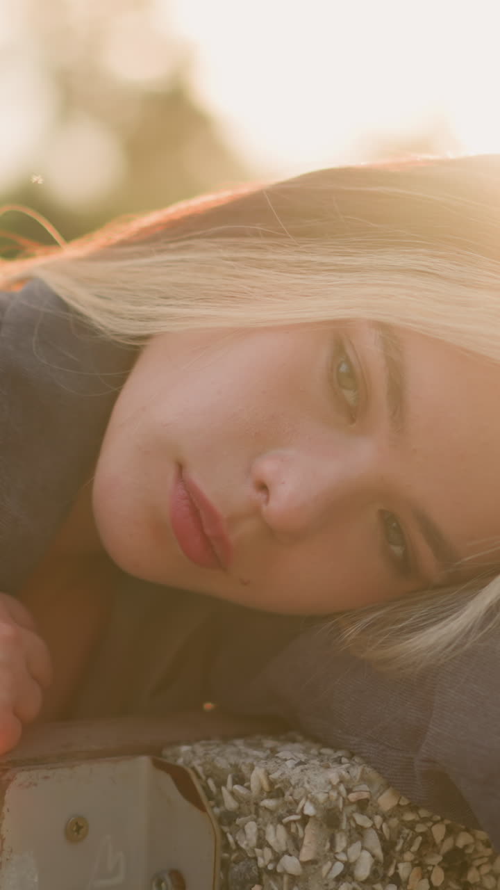 Woman seated on bench resting head on arm while gently touching hair, soft sunlight creating warm ambience around her, with a blur background