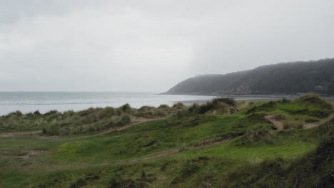 AERIAL:-Slow-sideways-pan-from-behind-grass-covered-sand-dune-looking-out-towards-overcast-Beach,-Oxwich,-4K-Drone