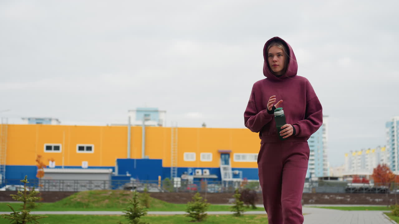 Jogger walking in burgundy tracksuit with hood up and white sneakers holding green water bottle on urban walkway lined with autumn shrubs and tall apartment buildings under overcast sky