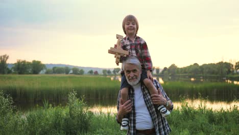 little happy boy sitting at the shoulders of his grandfather and playing with a wooden airplane toy in the park