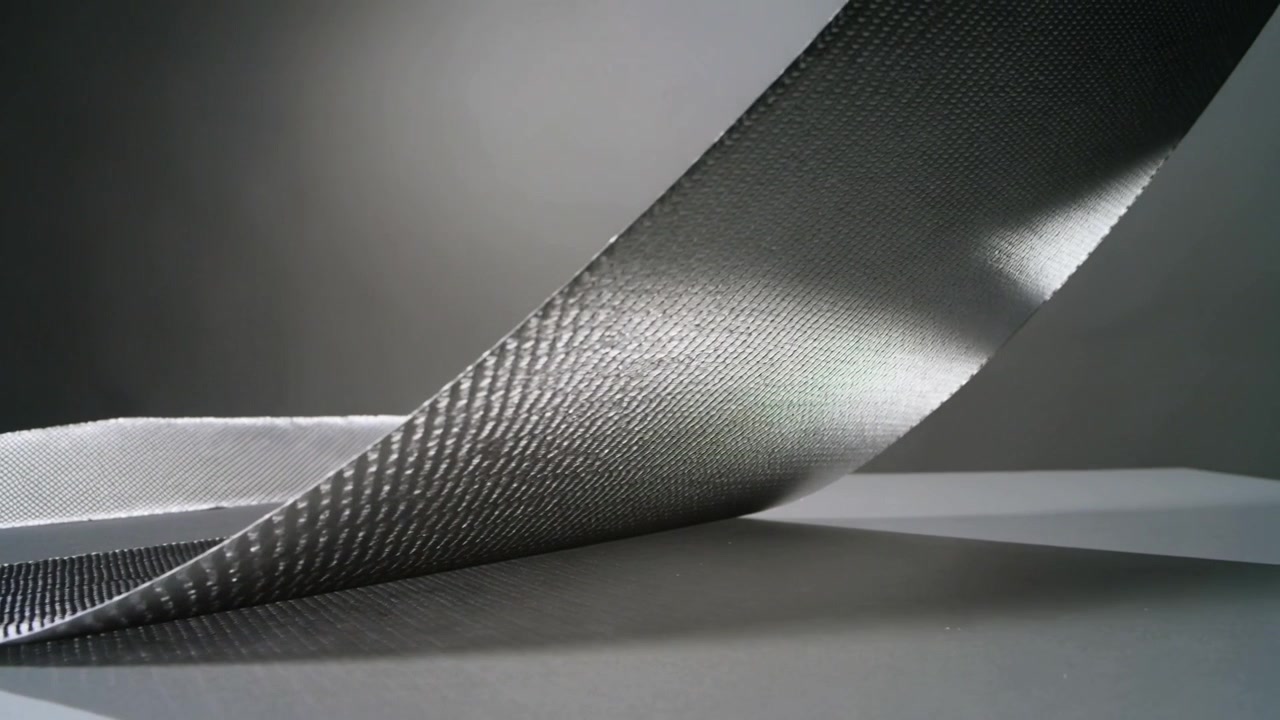 Lifting woven metal strip tilting across matte tabletop, showing glint and shadow on gray backdrop