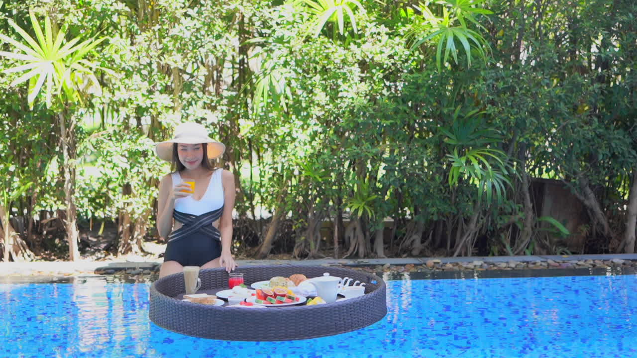 Static slow-motion shot of a lady grabbing a glass of orange juice from a floating pool tray with greenery in the background