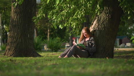 woman sitting outdoors with legs stretched, leaning against tree on grassy field, reading book under warm sunlight, tree leaves sway gently in breeze, background featuring a building and greenery