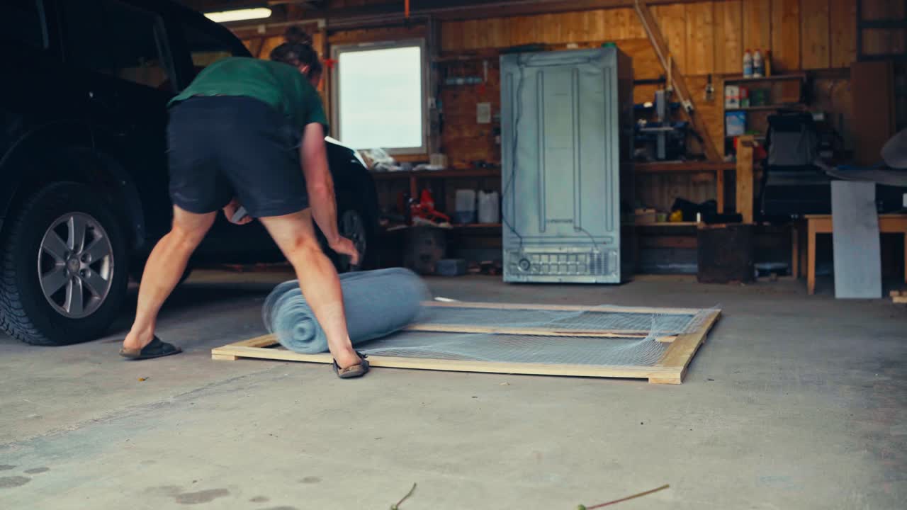 A Man Installing Wire Mesh In A Wooden Frame In A Garage. Static Shot