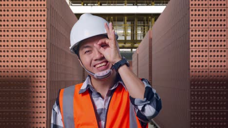 close up of asian male engineer with safety helmet showing ok hand sign over eye and smiling to camera while standing with red brick packed in stacks are stored