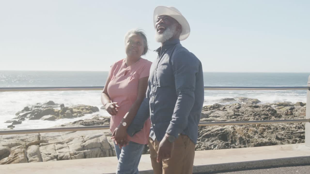 Happy senior african american couple walking along promenade by the sea, slow motion