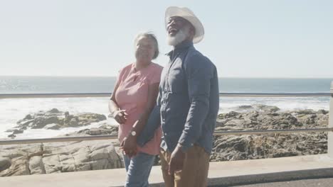 Happy-senior-african-american-couple-walking-along-promenade-by-the-sea,-slow-motion