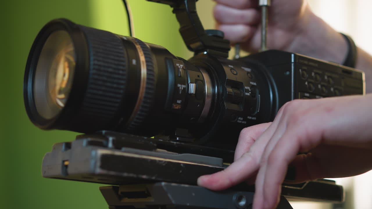 Close-up of camera mounted on tripod with lens in focus. Hand adjusting camera position. Green background and soft lighting create a cinematic, studio-like environment