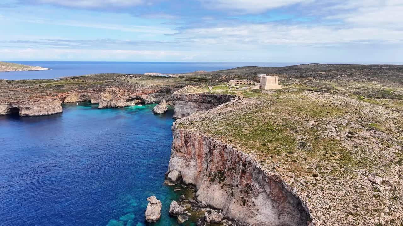 Veduta aerea di scogliere costiere rocciose e mare blu profondo vicino alla Torre di Santa Maria a Comino, Malta, con acqua limpida, grotte marine e una costa frastagliata sotto la luce del giorno intensa.