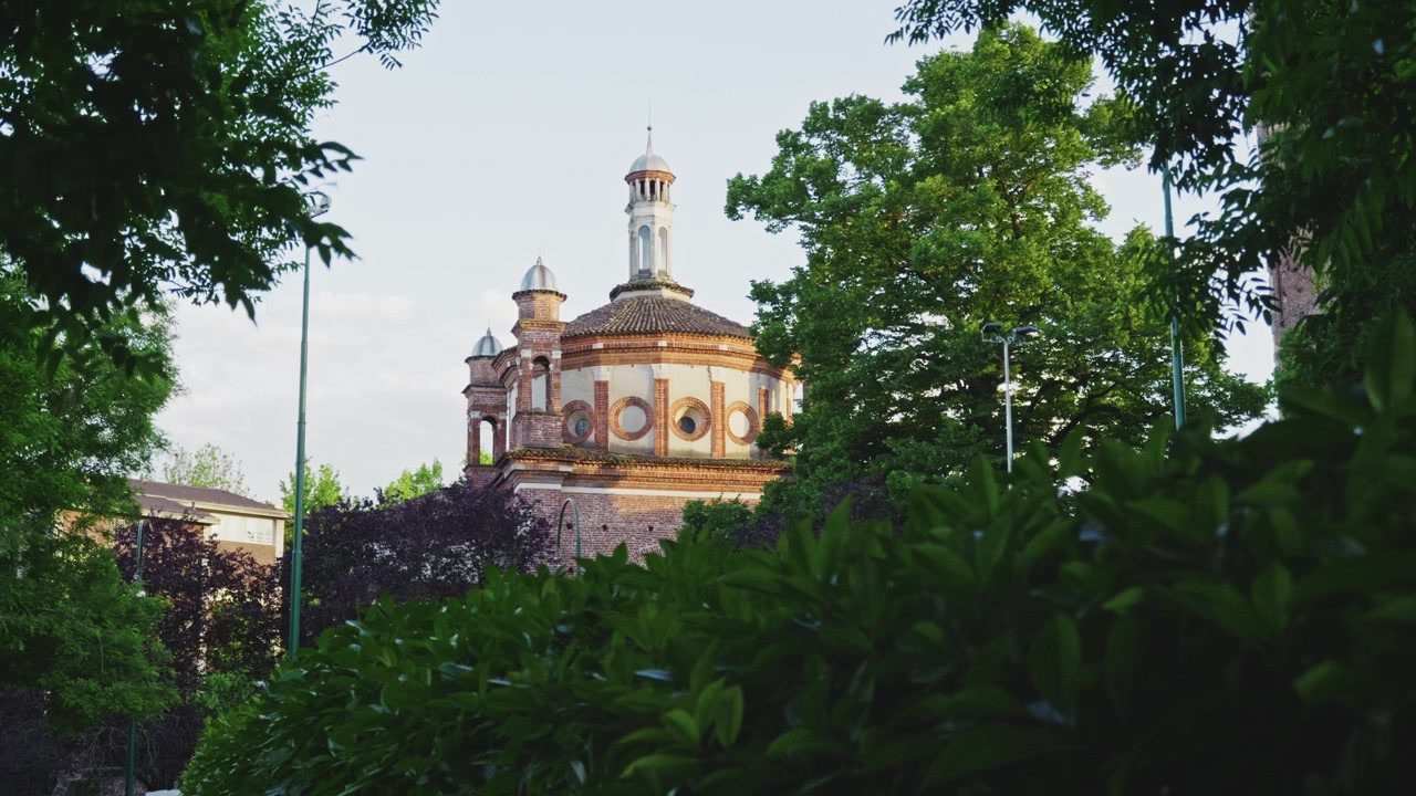 Uma bela igreja histórica parcialmente escondida por árvores verdes exuberantes, mesclando arquitetura e natureza no coração de Milão.