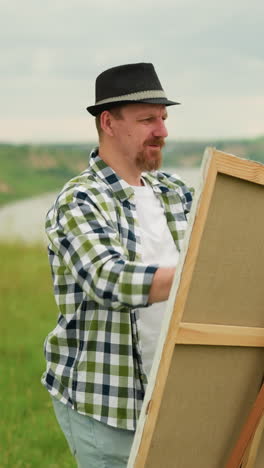 a painter wearing a black hat and plaid shirt is setting up a canvas on an easel in a grassy field. a woman in a white dress stands nearby, smiling as she watches the process