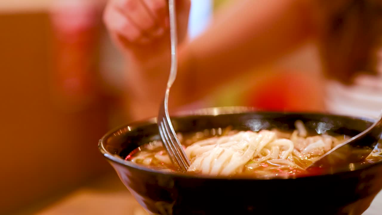 Close-up of noodles being lifted from a steaming bowl in a warmly lit Bangkok eatery