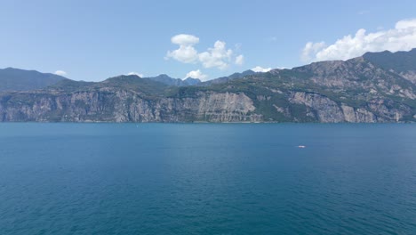 Una-Toma-Aérea-Panorámica-De-Las-Majestuosas-Montañas-Que-Se-Ciernen-Sobre-El-Lago-De-Garda-En-La-Encantadora-Ciudad-De-Malcesine,-Verona,-Italia-En-Un-Día-Pintoresco-Con-Un-Cielo-Azul-Claro-Y-Nubes-Esponjosas