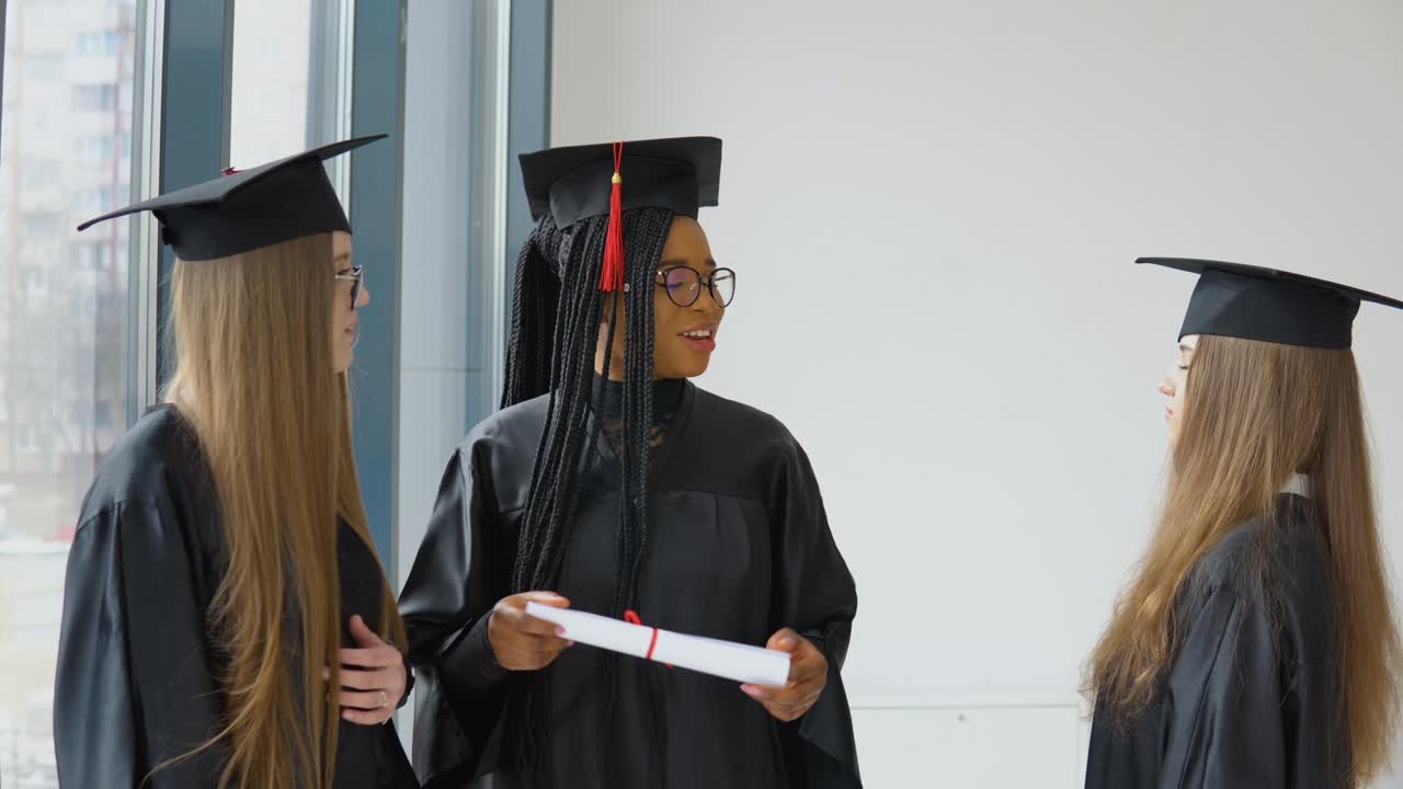 tres estudiantes femeninas de diferentes razas con un diploma en sus manos. graduados con túnicas negras y sombreros cuadrados tienen una conversación