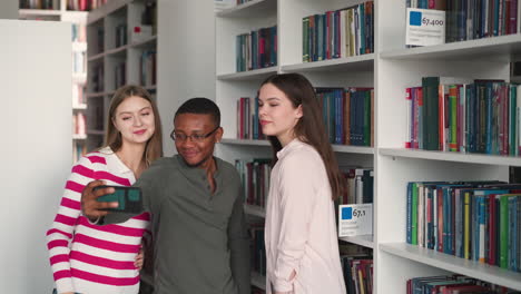 hombre negro con amigas se toma una selfie en la biblioteca. jóvenes positivos graban un video blog sobre el almacenamiento de literatura universitaria. amantes de las redes sociales