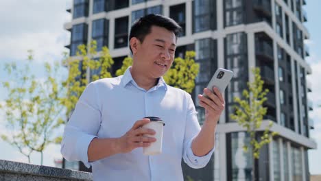 handsome smiling asian young adult entrepreneur business man browsing mobile phone surfing internet social media standing near beautiful modern building business district. successful chinese male.