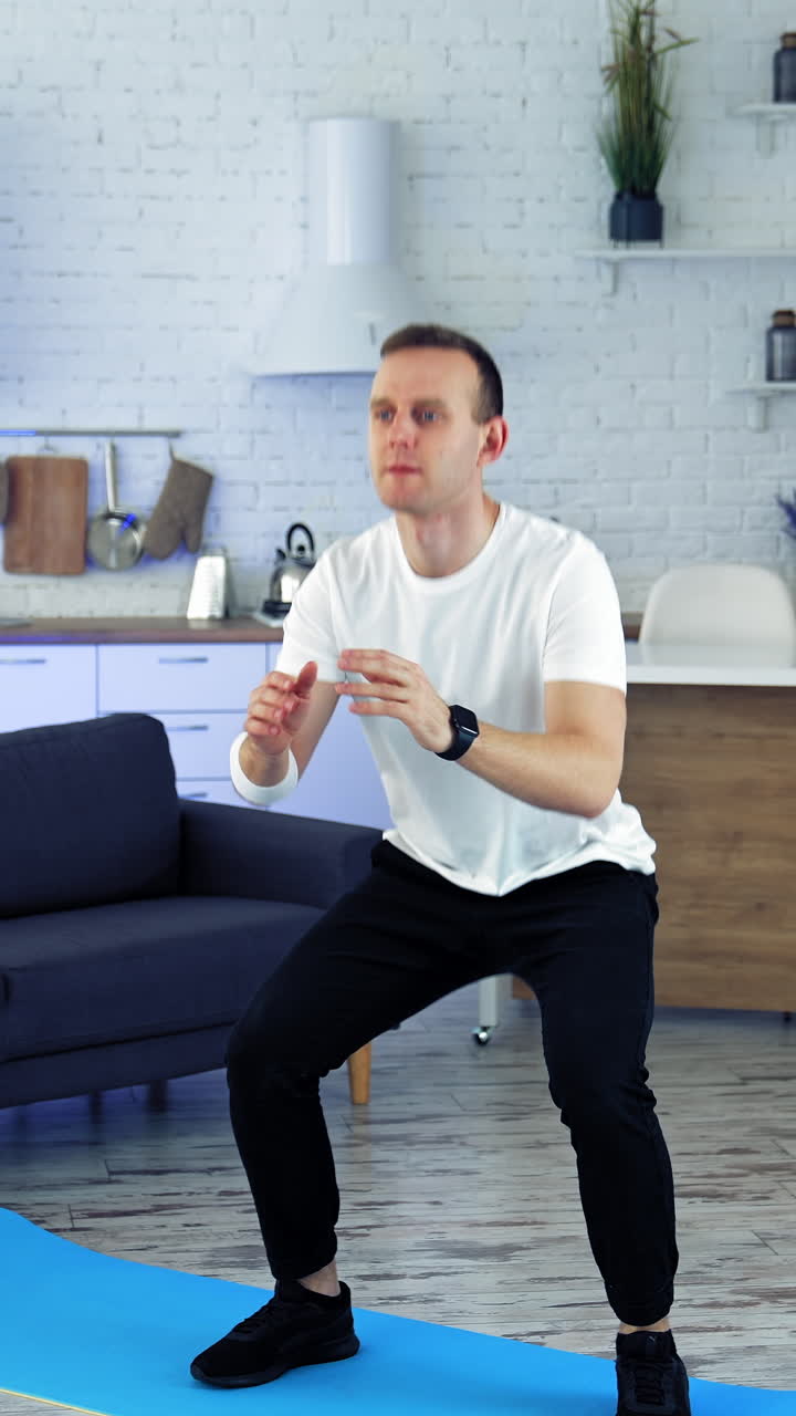 Home workout. Young man in white t-shirt and black pants doing squats exercises in kitchen interior. Sports training indoors. Slow motion. Vertical video