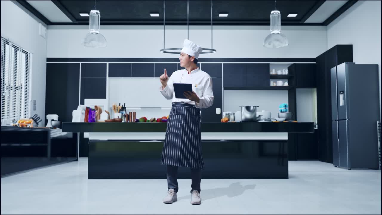 En asiatisk mandlig kok bruger en tablet og peger til siden, mens han står i et hjemmekøkken.