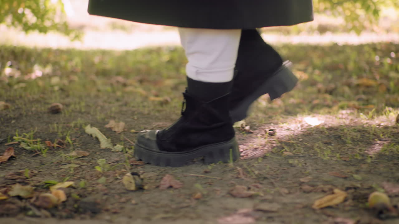 Leg view of park explorer in black boots strolling over leaf carpet on forest path, slow steps on soft ground, green canopy above, sunlit bokeh background, relaxed walk capturing movement