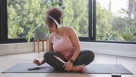 sitting on yoga mat, woman with headphones using smartphone in home gym