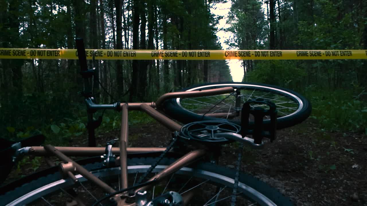 Low angle footage of a golden bicycle on a muddy forest lonely road with tall trees on the sides and a yellow Crime Scene Do Not Enter tape in the background hovering over the bicycle. Wheel spinning.