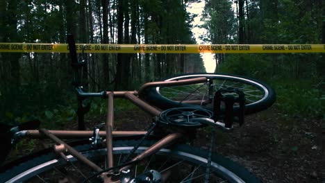 low angle footage of a golden bicycle on a muddy forest lonely road with tall trees on the sides and a yellow crime scene do not enter tape in the background hovering over the bicycle. wheel spinning.