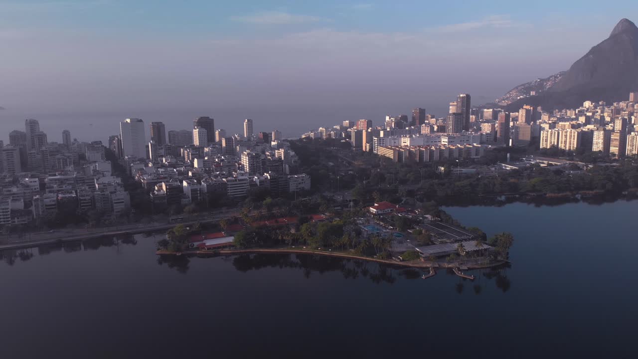 Aerial backward movement over the city lake towards island and club Caiçaras in Ipanema in the foreground and Leblon and Two Brothers mountain in the background at early morning sunrise