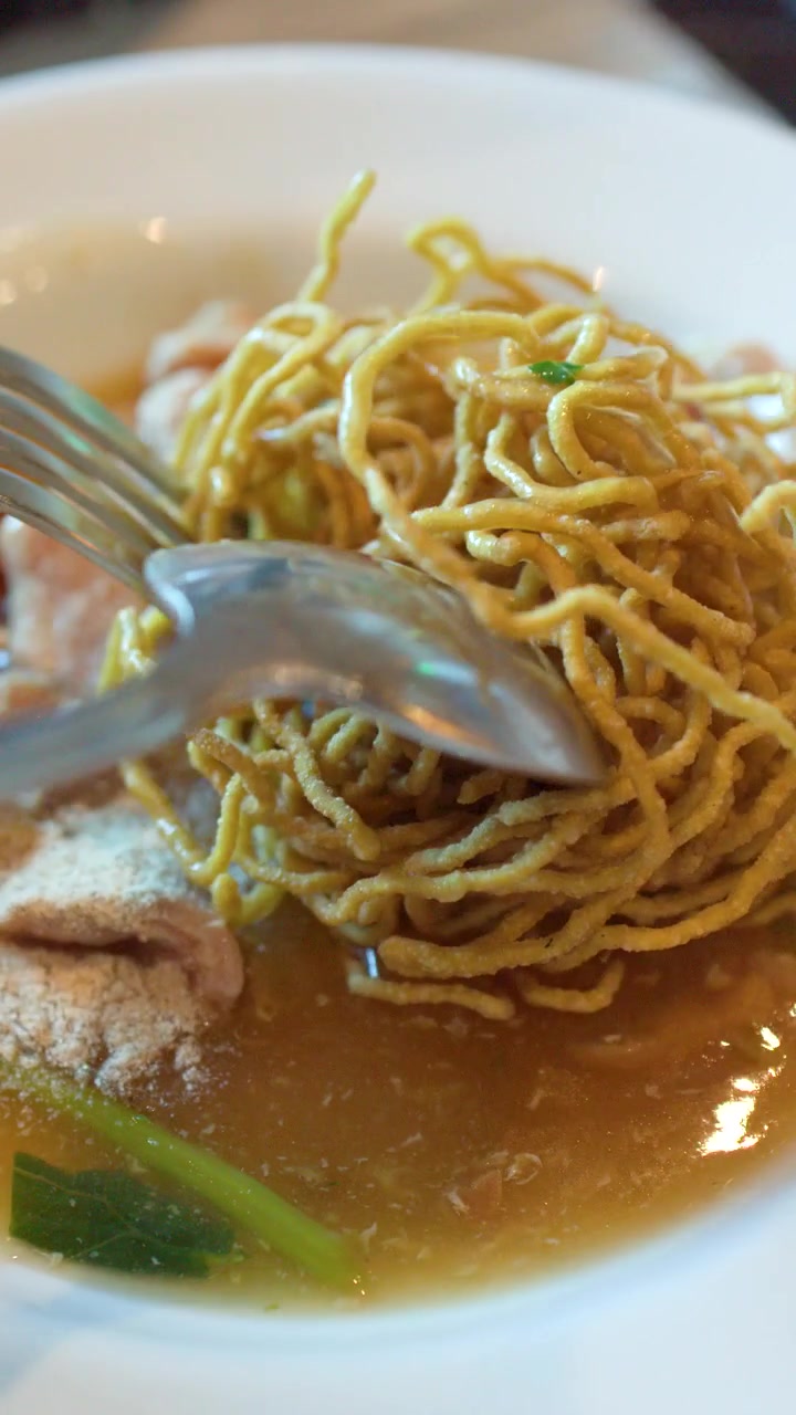Un primo piano mostra una forchetta e un cucchiaio che spezzano noodles croccanti in un sugo saporito.