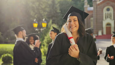 portrait of the happy young female graduate in a traditional clothes and cap smiling to the camera and holding her diploma on a sunny day