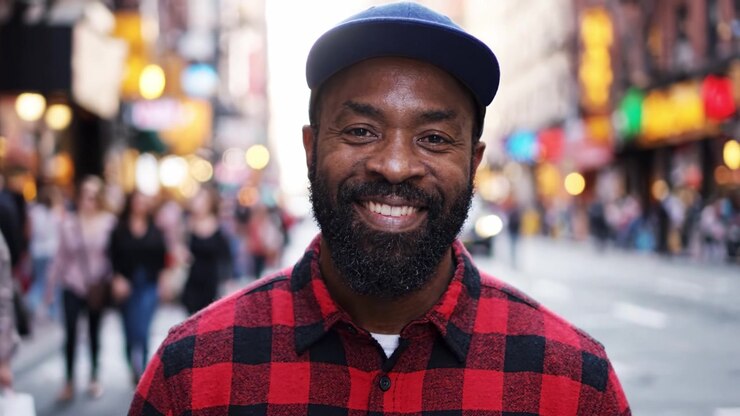 Un moment joyeux capturé en ville: un homme souriant avec une barbe, portant une casquette et une chemise à carreaux, au milieu d'un arrière-plan urbain animé