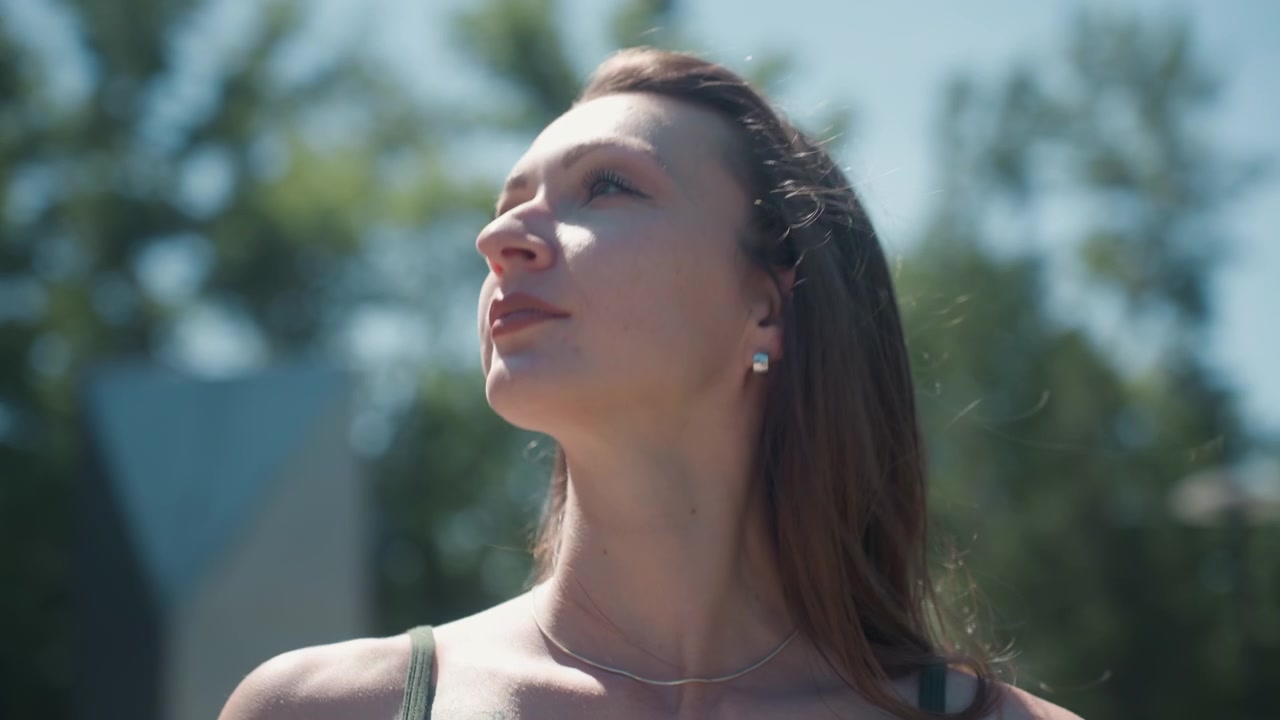 Primo piano di una giovane donna che si sistema gli occhiali da sole all'aperto sotto la luce solare intensa, rivelando dettagli come gioielli e la texture dei capelli. La scena presenta uno sfondo sfocato di vegetazione con luce naturale.