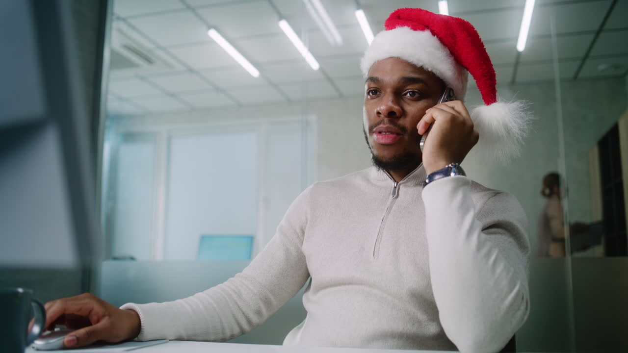 hombre con sombrero de santa trabajando en la computadora y hablando por teléfono en la oficina