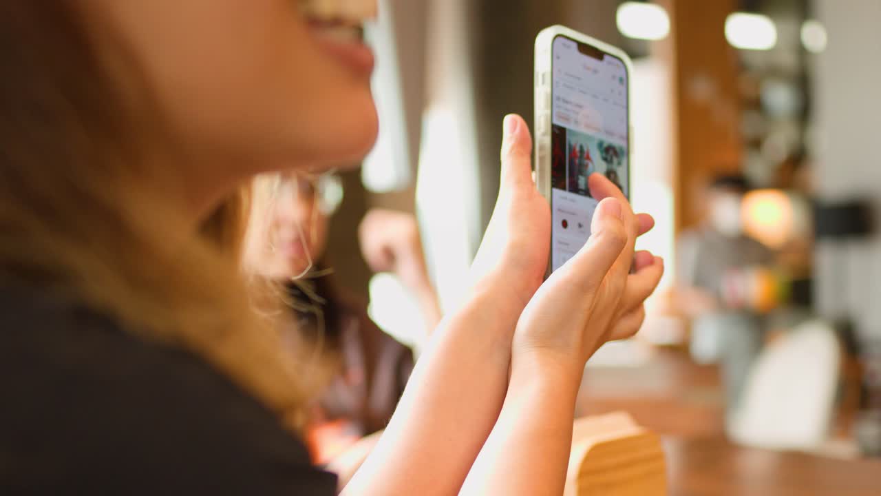 A woman holds a smartphone displaying a social media feed in a bright cafe setting