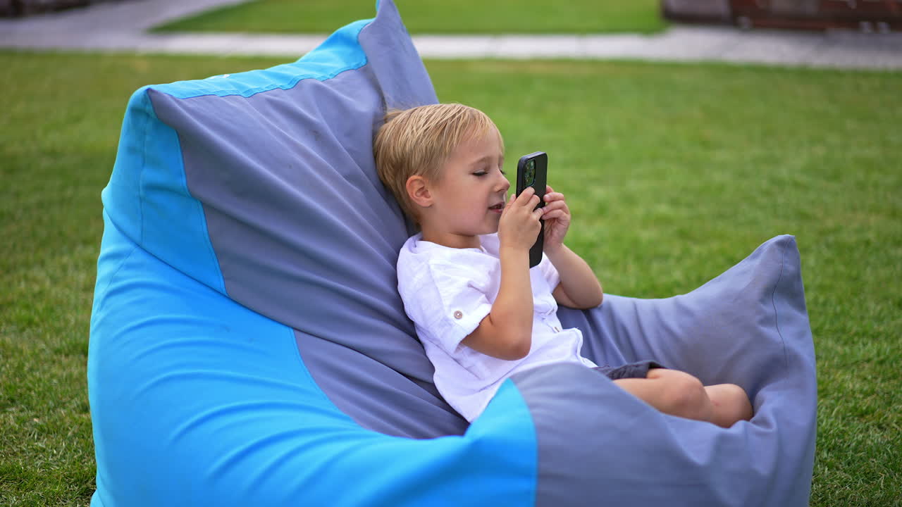Nice cute kid sitting outdoors in comfortable chair. Blond boy speaking on the smartphone on loud speaker. Green grass at backdrop.