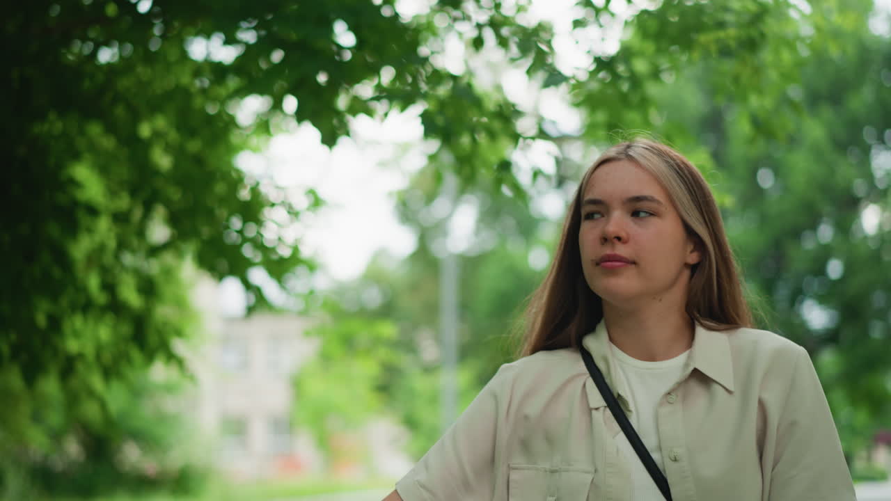 mujer joven con bolsa de cuerpo cruzado negra paseando al aire libre, rodeada de vegetación borrosa y luz solar suave filtrando a través de las hojas, con una vista de fondo de estructuras de construcción y árboles