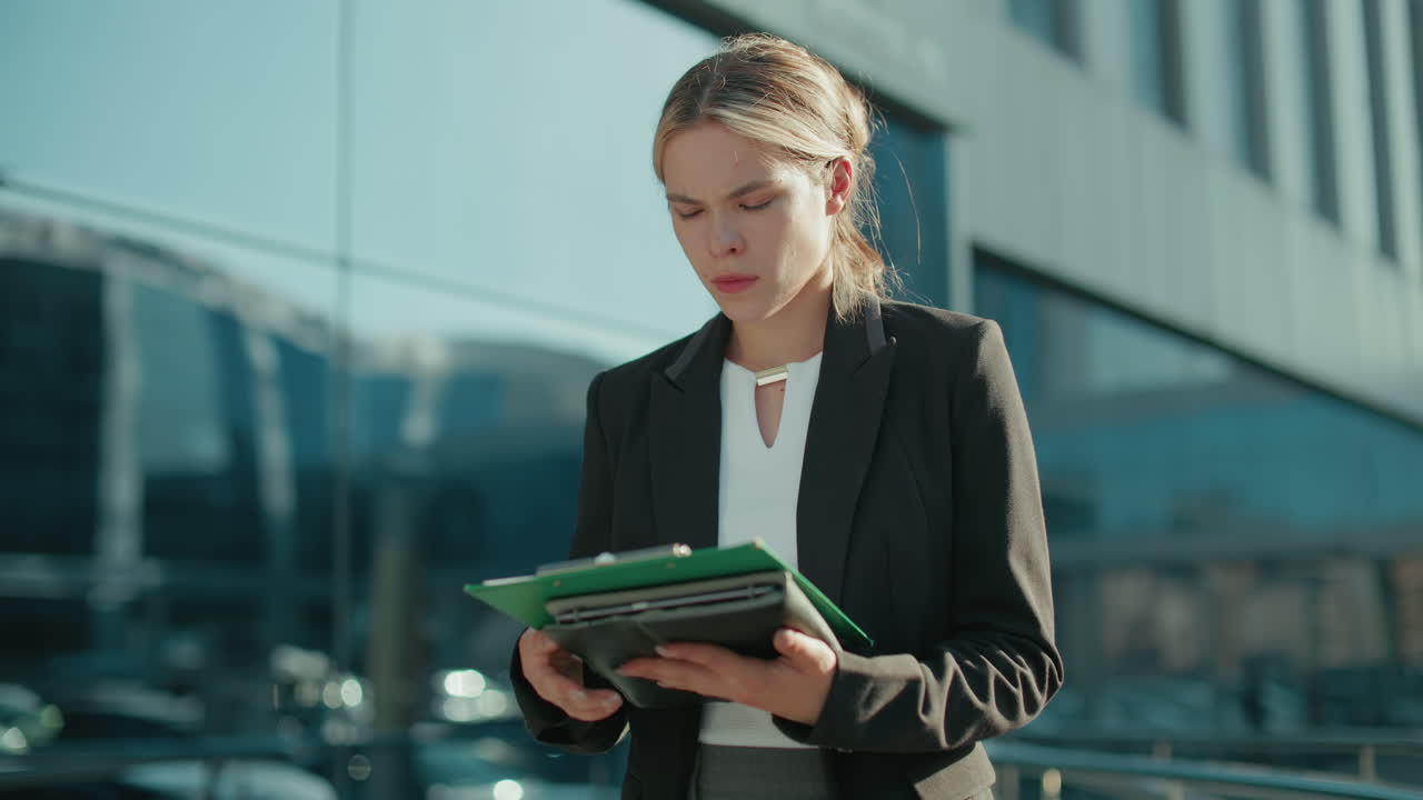 Business woman in black suit walking outdoors checking file in hand with focus and determination, reflected glass building and cars in blurred background