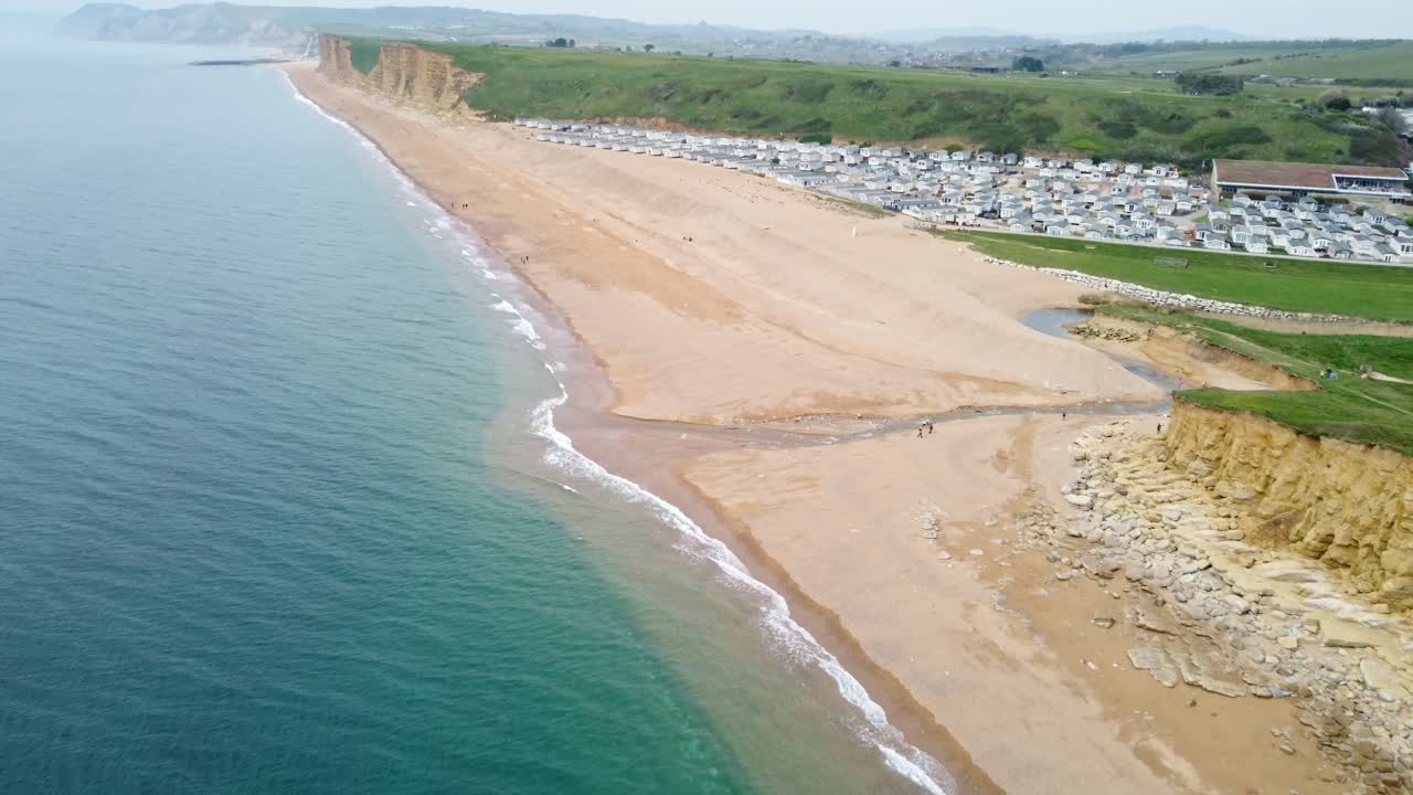 Aerial footage over a Freshwater Holiday Park beach front on the Jurassic Coast, Dorset. Shot is high looking down over the waves splashing on the sand before panning up showing some homes and cliffs.