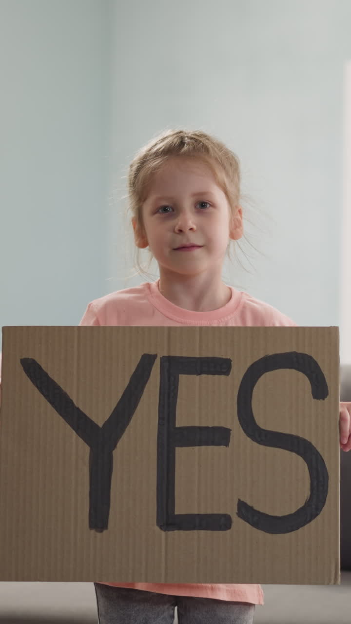 joyful, contented smiling little girl stands and holds poster with text YES in her hands, expresses her consent in room at home
