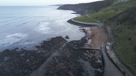 Nach-Unten-Gerichtete-Luftaufnahme-Eines-Leeren-Flussufers-Mit-Schwarzen-Steinen-Darauf-Und-Einigen-Wellen,-Die-Sich-Der-Küste-Am-Abend-In-Scarborough,-England,-Nähern