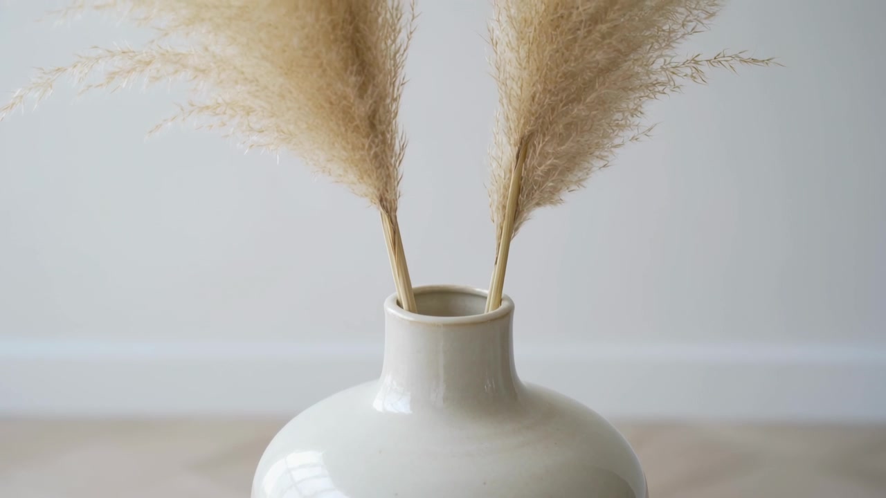 Piume di pampa che ondeggiano reagendo a una leggera corrente d'aria su un tavolo, vicino a un vaso di ceramica lucido bianco sporco.