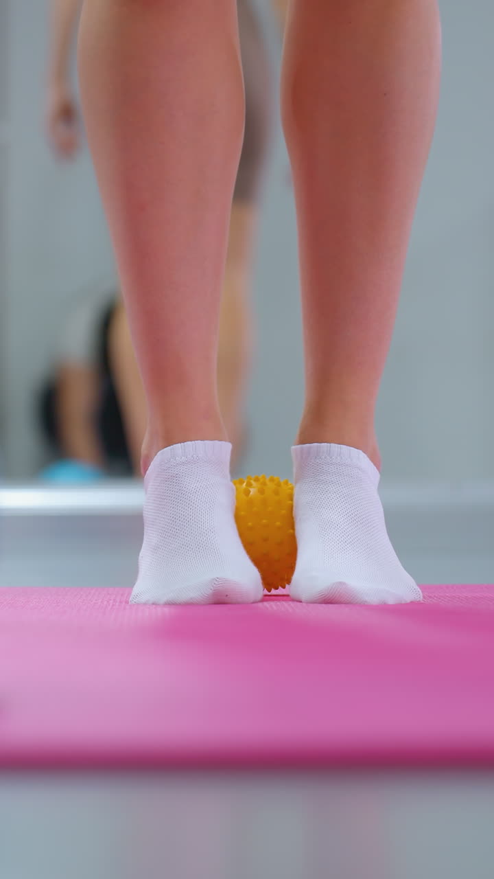 vista de la parte inferior de la pierna durante una sesión de entrenamiento con una pequeña pelota amarilla entre los tobillos y una pelota azul en la parte delantera, un gran espejo que refleja a otros participantes en el fondo